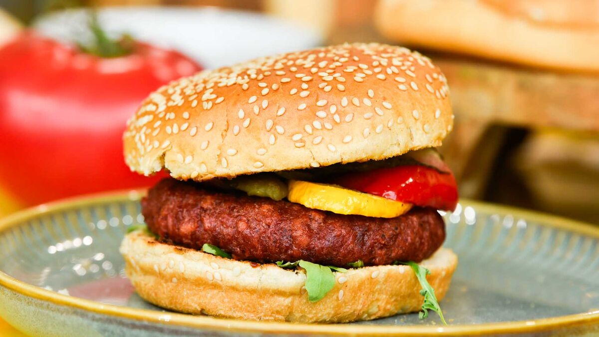 Roślinne zamienniki mięsa na grillu - burger a'la wołowy. Burger z sezamową bułką podano na talerzu. W środku znajduje się roślinny kotlet, rukola, grillowana papryka i żółte warzywo.