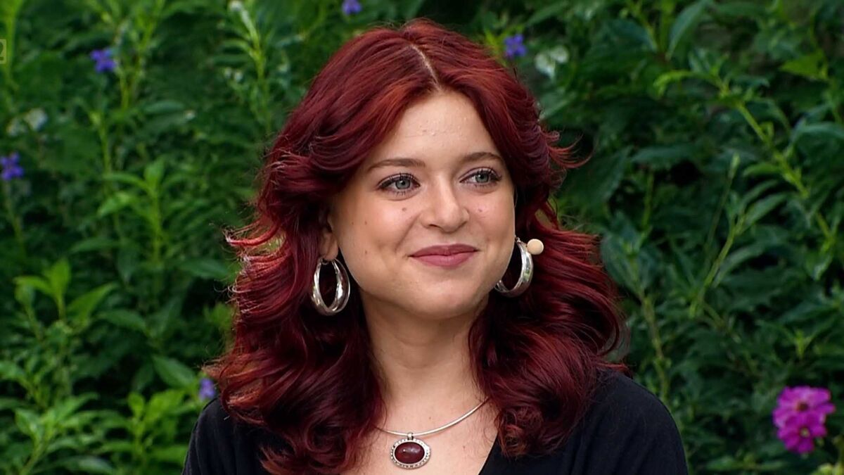 Gabriela Raczyńska siedzi na kanapie w plenerowym studiu telewizyjnym. Ma na sobie czarną bluzkę, srebrne kolczyki i naszyjnik z ciemnym kamieniem oraz mikrofon nagłowny przy policzku. W tle widoczne są gęste zielone rośliny i kwitnące kwiaty.