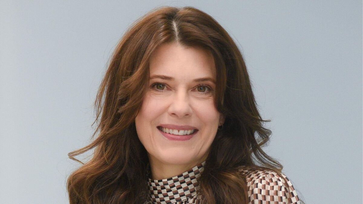 Joanna Sydor ma długie, brązowe włosy ułożone w luźne fale i patrzy przed siebie. Ma na sobie bluzkę z wysokim kołnierzem w drobną biało-brązową kratę. W tle znajduje się jednolita, jasnoszara powierzchnia.