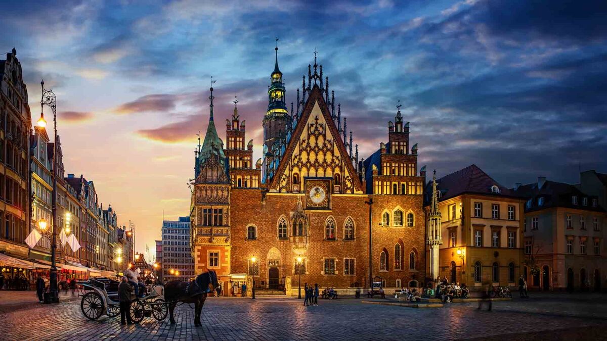 Ratusz na wrocławskim Rynku oświetlony wieczornym światłem, z widocznym zegarem na fasadzie. Na placu stoją kamienice oraz zaparkowana dorożka z koniem.