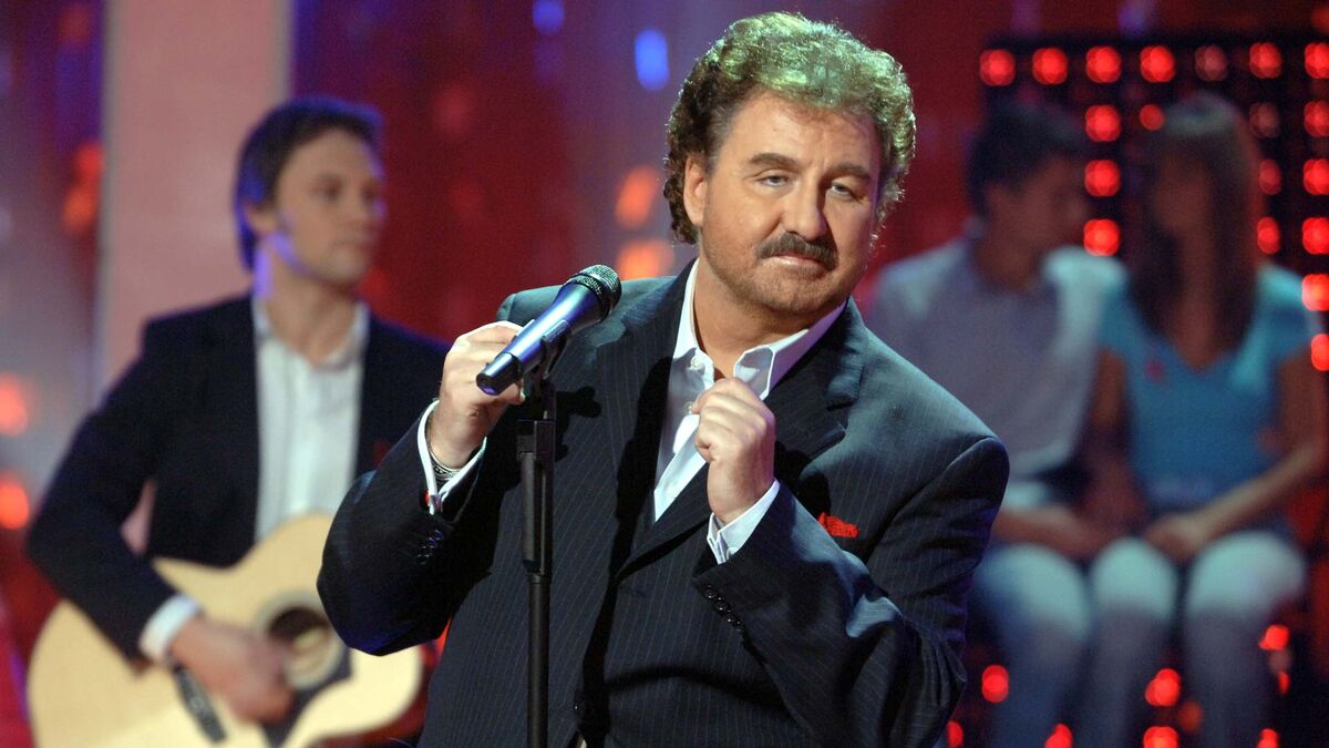 Krzysztof Krawczyk stoi przy mikrofonie, ubrany w ciemny garnitur i białą koszulę. W jednej dłoni trzyma mikrofon, a drugą ma uniesioną przed sobą. W tle widać gitarzystę, siedzące osoby oraz czerwone elementy oświetlenia scenicznego.