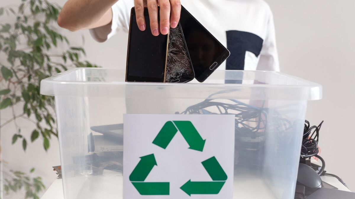 Osoba wrzuca kilka starych smartfonów, w tym jeden z pękniętym ekranem, do przezroczystego pojemnika przeznaczonego na elektroodpady. Na przodzie pojemnika widnieje duży zielony symbol recyklingu, a w środku widać też kable i inne części elektroniczne. Jasne tło i stojąca obok roślina doniczkowa podkreślają ideę dbania o środowisko poprzez odpowiednią utylizację zużytego sprzętu.