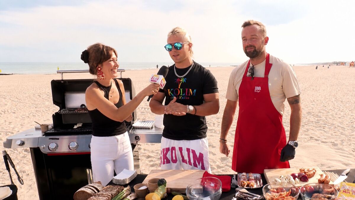 Skolim stoi na plaży i udziela wywiadu kobiecie trzymającej mikrofon, a obok nich stoi mężczyzna w czerwonym fartuchu. Skolim ma na sobie czarną koszulkę, białe spodenki i okulary przeciwsłoneczne. W tle widać grill, piaszczystą plażę, morze oraz nieliczne osoby spacerujące w oddali.