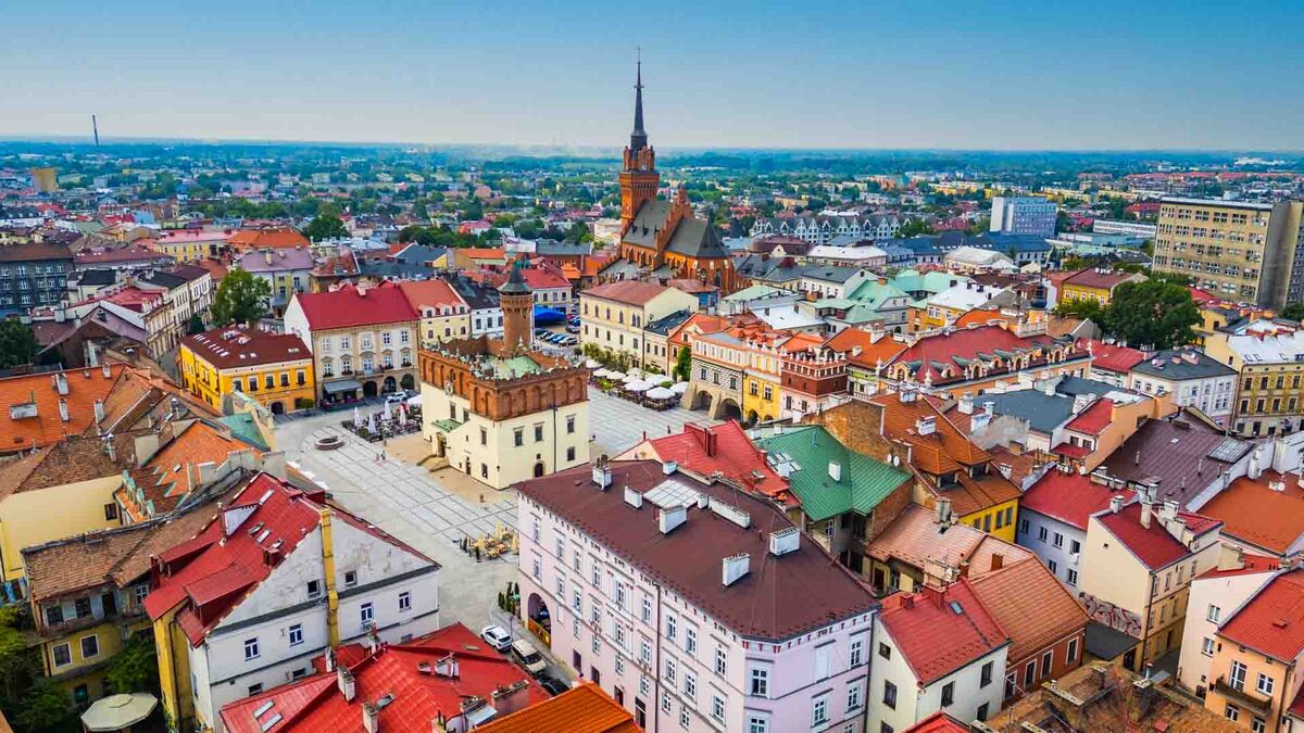 Widok z góry na centrum Tarnowa obejmuje rynek i zwartą zabudowę z kolorowymi dachami. Pośród kamienic widoczny jest ratusz z wieżą, a dalej wznosi się wysoka wieża kościoła. W tle rozciągają się kolejne dzielnice miasta oraz pas zieleni na horyzoncie.