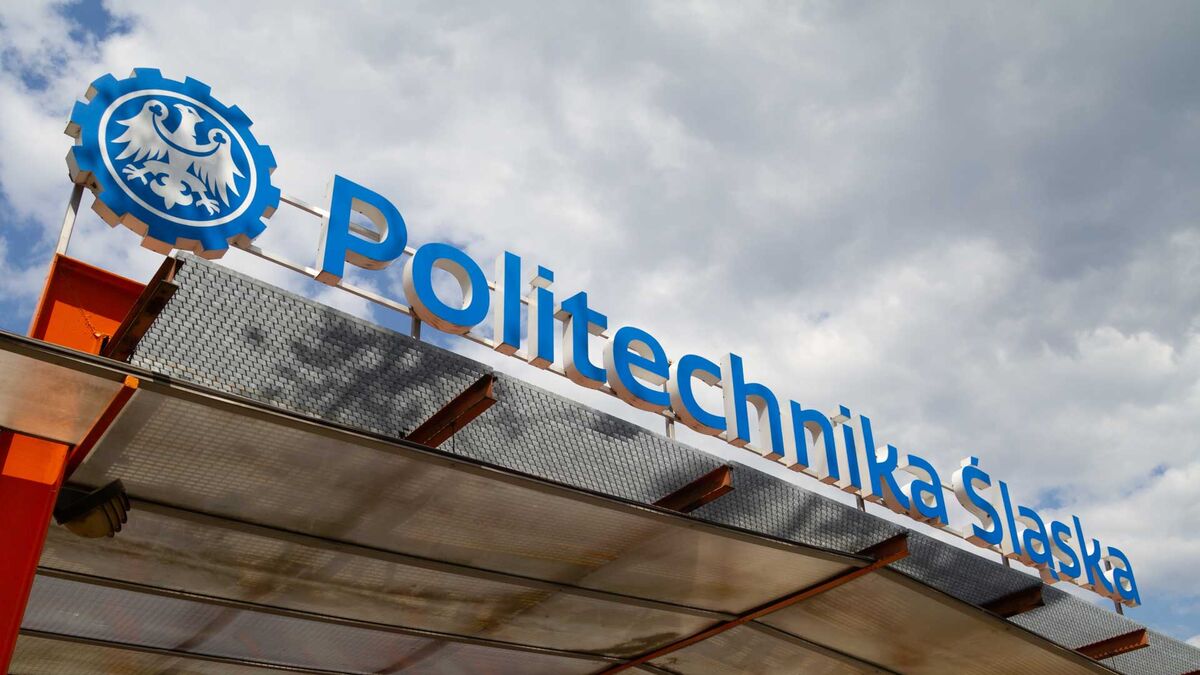 Politechnika Śląska – widoczny znak uczelni na dachu budynku. Na pierwszym planie znajduje się niebieskie godło w kształcie koła z orłem. W tle widać chmury na niebie oraz konstrukcję zadaszenia.