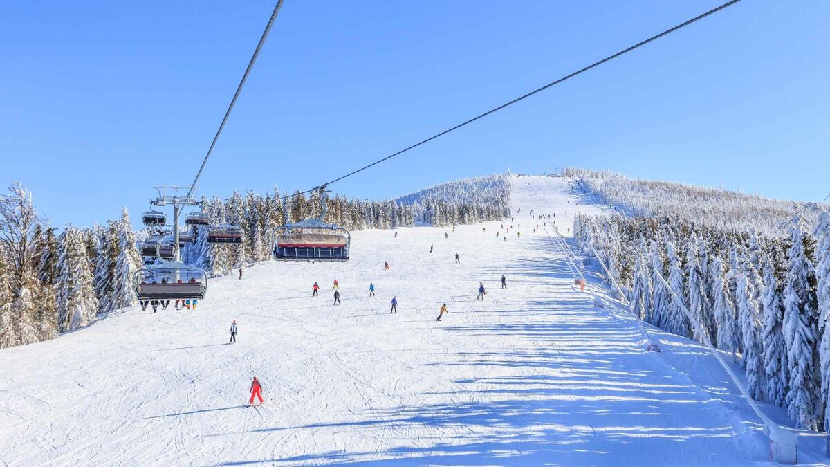 Szeroka, ośnieżona trasa narciarska wypełnia kadr, a nad stokiem sunie wyciąg krzesełkowy. Na białym zboczu widać wielu narciarzy i snowboardzistów, a po bokach stoją gęste, oszronione drzewa. Bezchmurne niebo i mocne słońce rzucają długie cienie, podkreślając zimowy, pogodny klimat.