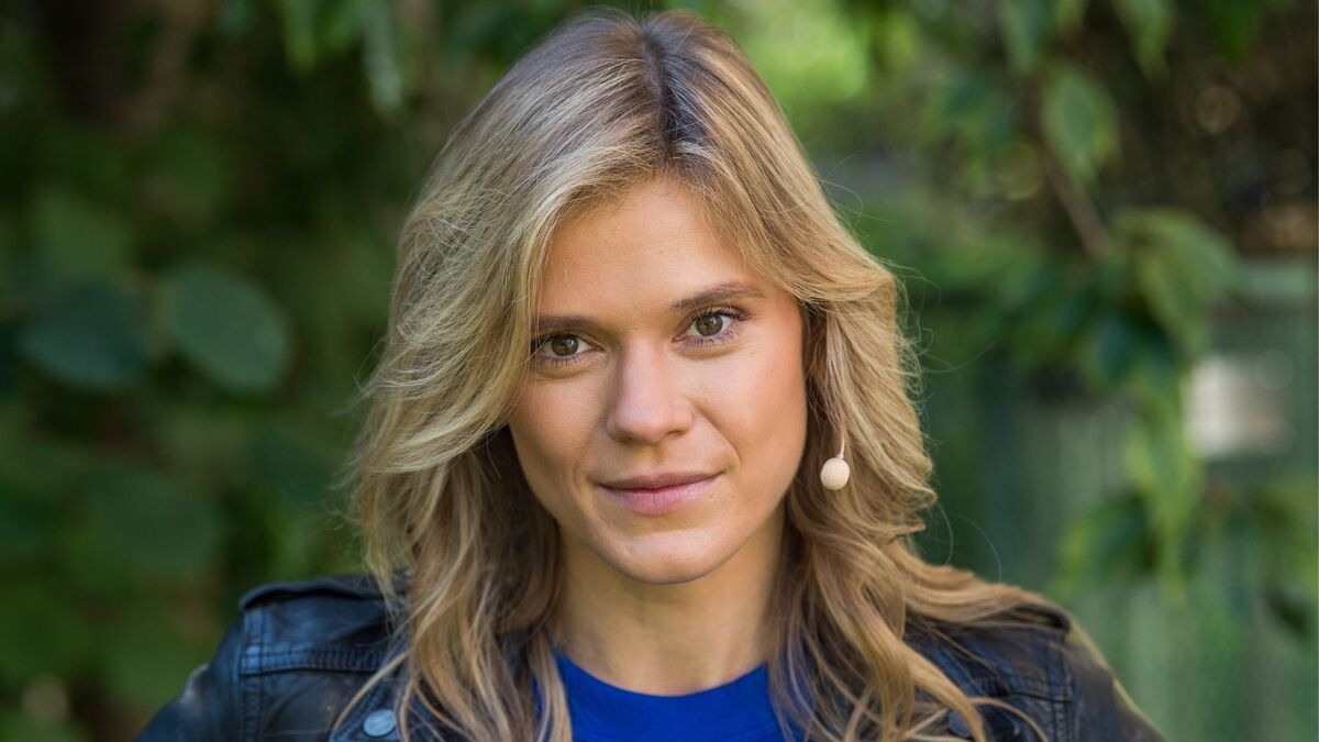 Barbara Liberek ma rozpuszczone blond włosy i jest ubrana w niebieską bluzkę oraz czarną skórzaną kurtkę. Przy twarzy widoczny jest niewielki mikrofon nagłowny. W tle znajdują się rozmyte zielone liście i roślinność.