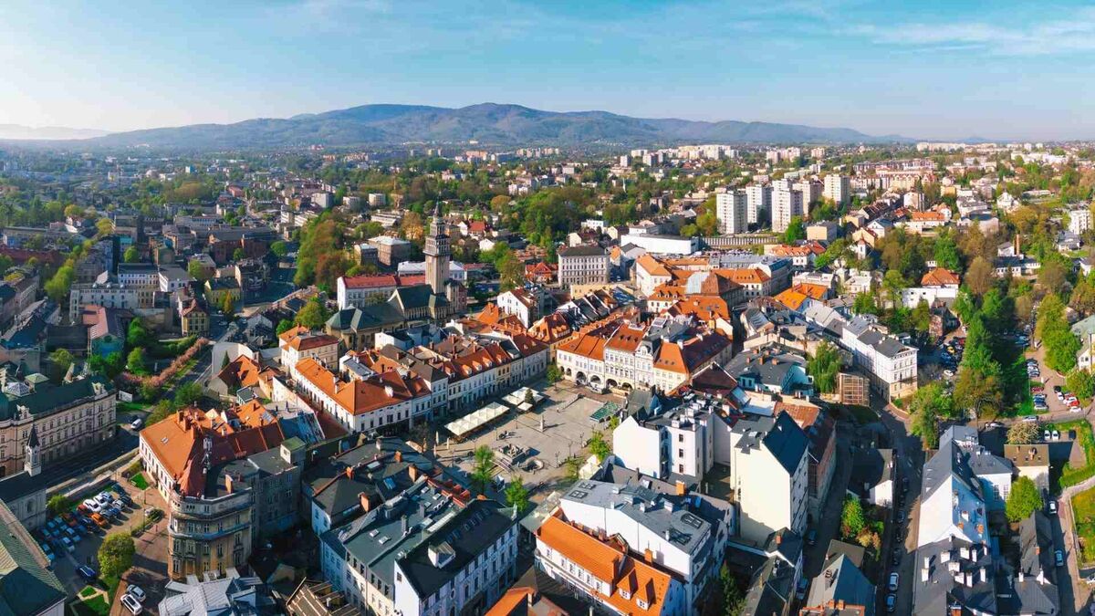 Panoramiczny widok na Bielsko-Białą z lotu ptaka. W centrum miasta widoczny jest rynek otoczony zabytkowymi kamienicami z charakterystycznymi czerwonymi dachami oraz wieża ratusza górująca nad okoliczną zabudową. W tle rozciągają się zielone wzgórza Beskidów, a między budynkami widoczne są liczne drzewa i tereny zielone, podkreślające malowniczy charakter miasta.