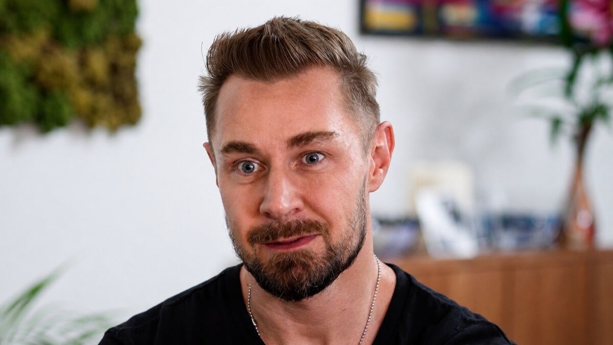 Patryk Kwiecień patrzy w obiektyw z lekko uniesionymi brwiami i subtelnym uśmiechem. Ma krótkie włosy, zarost oraz czarną koszulkę, a na szyi widoczny jest cienki łańcuszek. W tle znajdują się rozmyte elementy wnętrza, w tym rośliny i drewniana komoda.